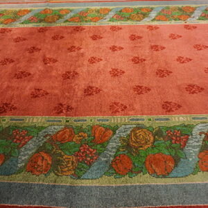 Rose-colored rug with small darker motifs in the field and broad floral borders featuring orange, green, and blue details.