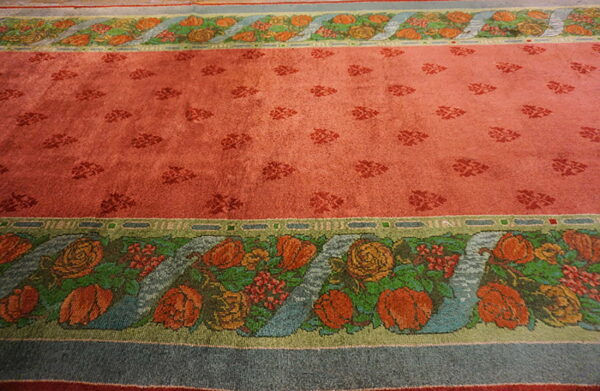 Rose-colored rug with small darker motifs in the field and broad floral borders featuring orange, green, and blue details.