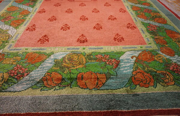 Close-up of a rug with a salmon pink field and pictorial borders of orange flowers, green leaves, and blue water.