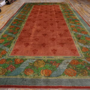 Reddish-orange rug with floral medallions and a wide border of colorful roses rests on distressed wood flooring.