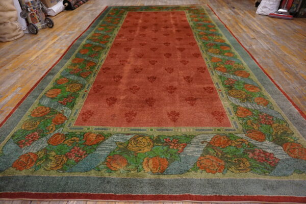 Reddish-orange rug with floral medallions and a wide border of colorful roses rests on distressed wood flooring.