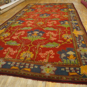 A deep red rug featuring multicolored abstract floral patterns lies on a worn light wood floor.