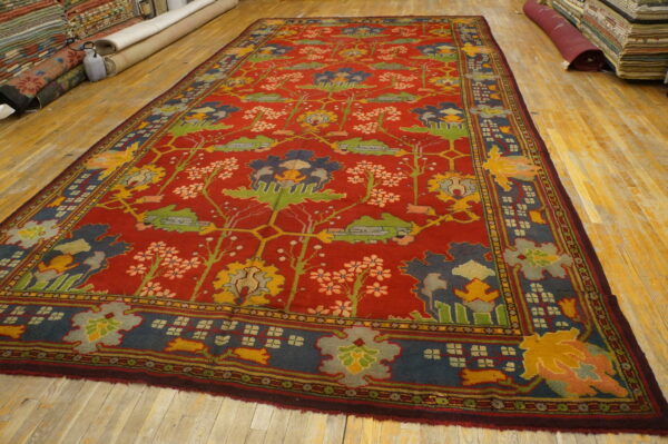 A deep red rug featuring multicolored abstract floral patterns lies on a worn light wood floor.