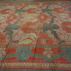 A large, faded rug with a traditional floral design in reddish-orange, green, and tan on wooden planks.