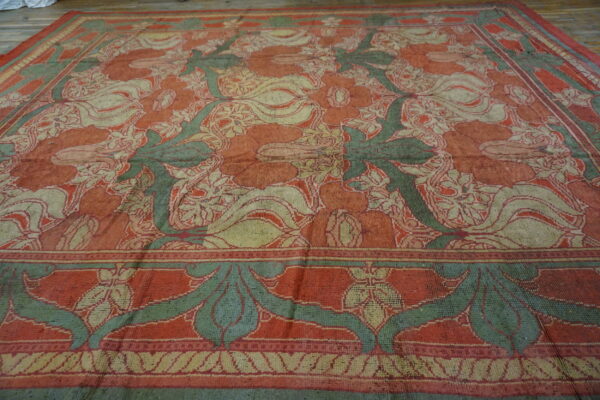 A large, faded rug with a traditional floral design in reddish-orange, green, and tan on wooden planks.