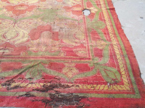 Distressed red and olive green floral rug on concrete with a large hole and noticeable thread unraveling.