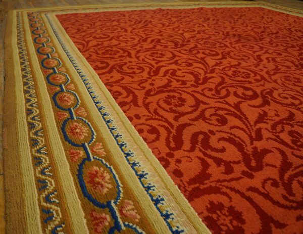 Terra cotta rug with damask field and colorful blue and gold border rests on hardwood floor.
