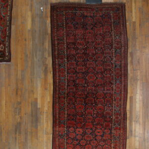 Dark red rug with geometric blue patterns rests on light wooden floorboards with other rugs visible.