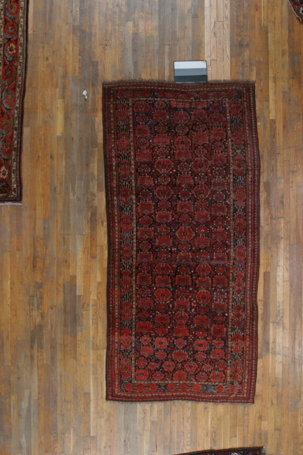 Dark red rug with geometric blue patterns rests on light wooden floorboards with other rugs visible.