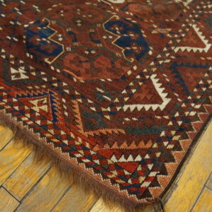 Corner of a deep red rug displaying complex blue, white, and rust geometric motifs on wide, aged wood flooring.