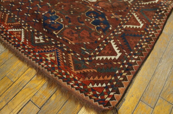 Corner of a deep red rug displaying complex blue, white, and rust geometric motifs on wide, aged wood flooring.