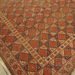 Rust-red rug with diagonal geometric shapes in blue and tan lies on distressed gray wood floorboards.