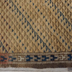 Textured tan rug close-up featuring blue geometric diamonds and a stepped border over light wood planks.