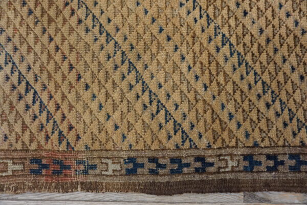 Textured tan rug close-up featuring blue geometric diamonds and a stepped border over light wood planks.