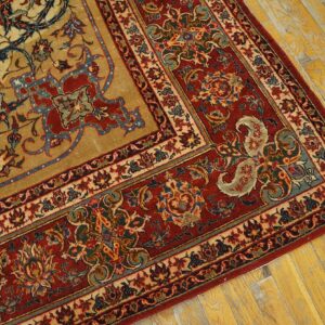 Close-up of a traditional gold and tan rug with red, blue, and cream floral borders on distressed yellow wood flooring.