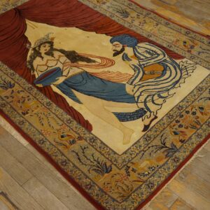 Figurative rug displaying a scene with two figures and red drapery, framed by a blue and tan floral border on wood flooring.
