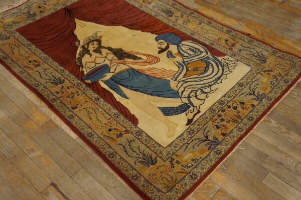 Figurative rug displaying a scene with two figures and red drapery, framed by a blue and tan floral border on wood flooring.