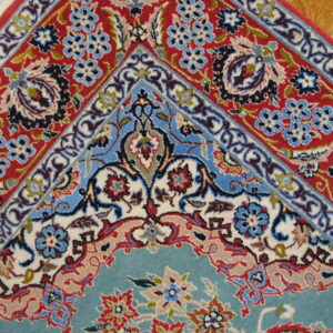 Intricate floral rug corner featuring a teal center, red border, blue details, and white fringe on wood flooring.