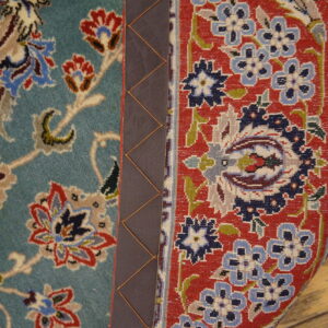 Two contrasting floral rug sections, teal and red, meet at a dark stitched binding over wooden planks.