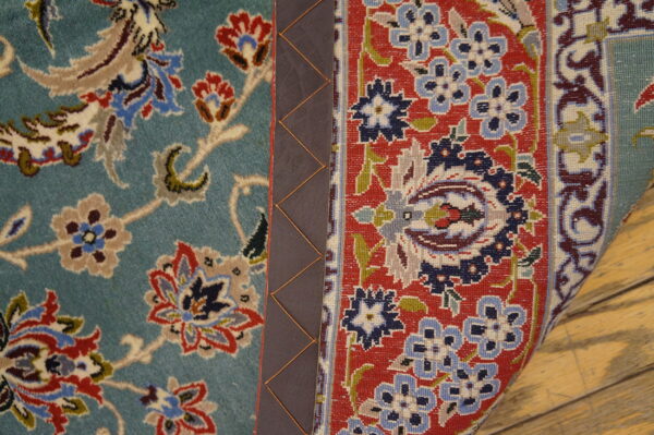 Two contrasting floral rug sections, teal and red, meet at a dark stitched binding over wooden planks.