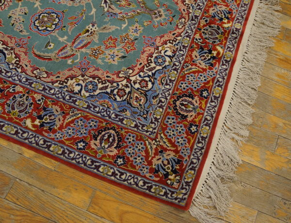 Teal and red patterned rug corner with fringe rests on light brown distressed wood flooring.