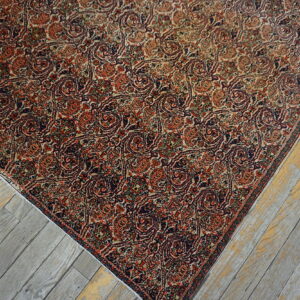 The corner of a dense, reddish-orange and navy traditional patterned rug rests on faded gray plank flooring.