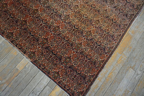 The corner of a dense, reddish-orange and navy traditional patterned rug rests on faded gray plank flooring.