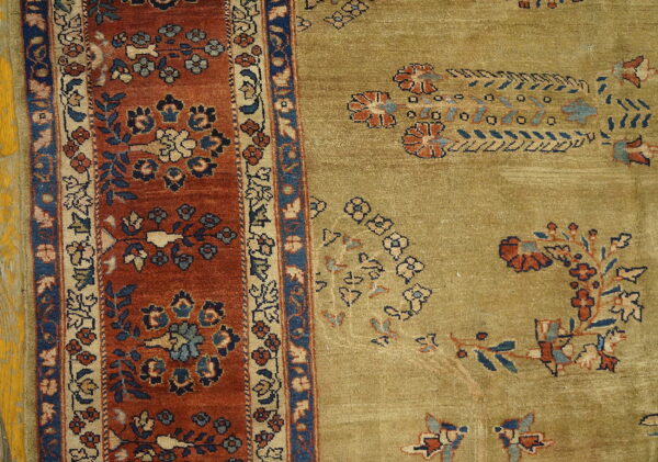 Detail of an olive-green rug field next to a rust and blue floral border on distressed yellow flooring.