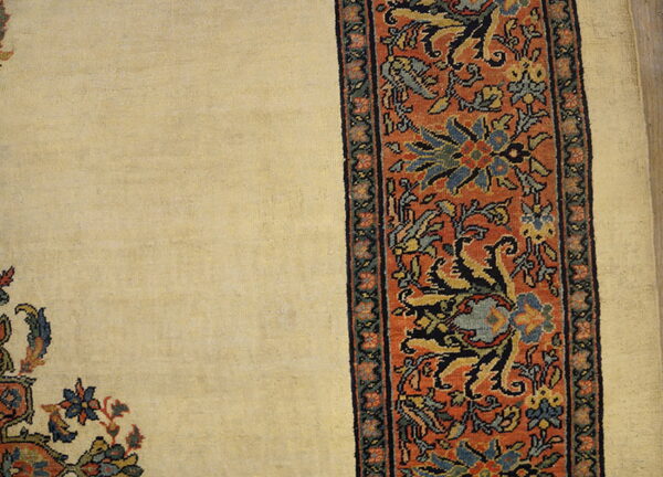 Cream rug field detail alongside a wide, rusty-orange border featuring traditional blue and yellow floral motifs.