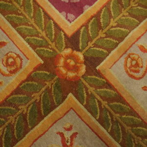 Textured rug close-up shows green leaves, an orange flower, gold stripes, and grey and magenta background panels.