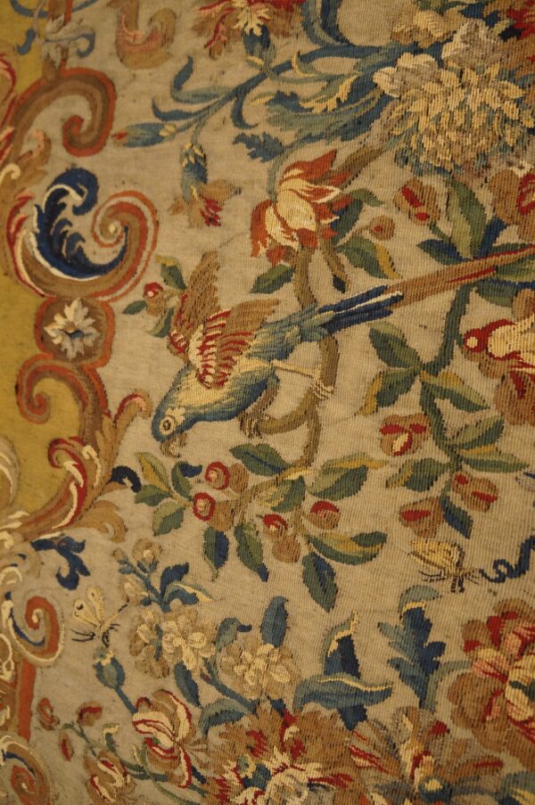 Muted flatweave detail with a parrot, red roses, and scrolling blue patterns on a pale tan ground.