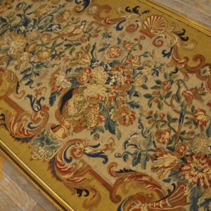 Muted, framed tapestry shows dense floral and bird patterns in gold, blue, and red over wood planks.