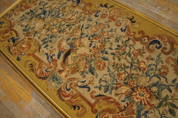 Muted, framed tapestry shows dense floral and bird patterns in gold, blue, and red over wood planks.