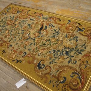 Ornate flat-weave tapestry featuring faded floral designs and a wide mustard scroll border, framed in gold on plank flooring.