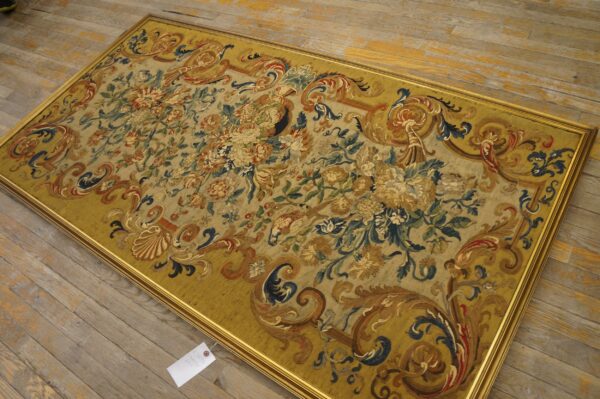 Ornate flat-weave tapestry featuring faded floral designs and a wide mustard scroll border, framed in gold on plank flooring.