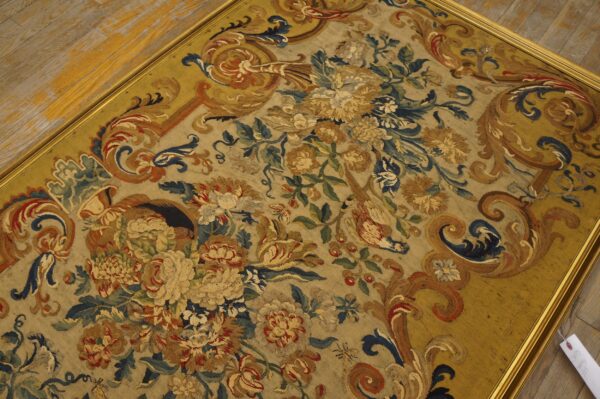 Ornate floral flatweave textile in gold, blue, and tan framed by a thin metallic border on wood.