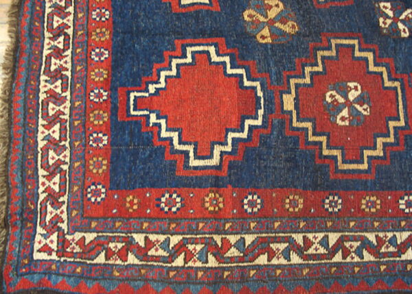 Low-pile blue, red, and cream rug corner displaying intricate geometric guls and borders with fringe.