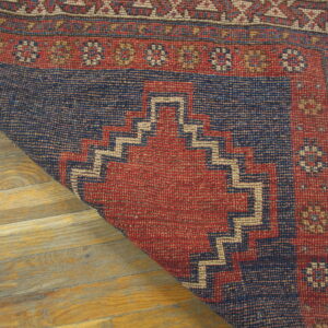Red and dark blue geometric rug with stepped patterns partially rolled back on light wood flooring.