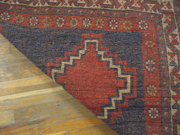 Red and dark blue geometric rug with stepped patterns partially rolled back on light wood flooring.