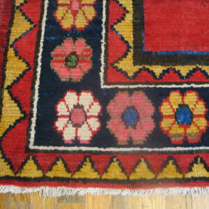 Red and navy rug with geometric floral motifs and jagged yellow borders resting on hardwood flooring.