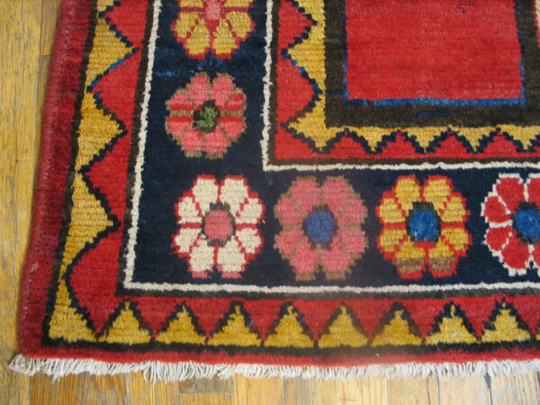 Red and navy rug with geometric floral motifs and jagged yellow borders resting on hardwood flooring.