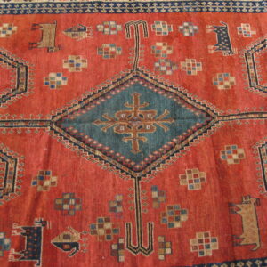 Red-orange tribal rug with a low pile, teal diamond medallion, and stylized animal figures.