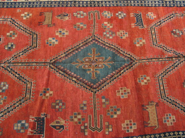 Red-orange tribal rug with a low pile, teal diamond medallion, and stylized animal figures.