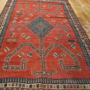 Rusty red tribal rug with a geometric pattern, central teal diamond medallion, and navy borders on wood floor.