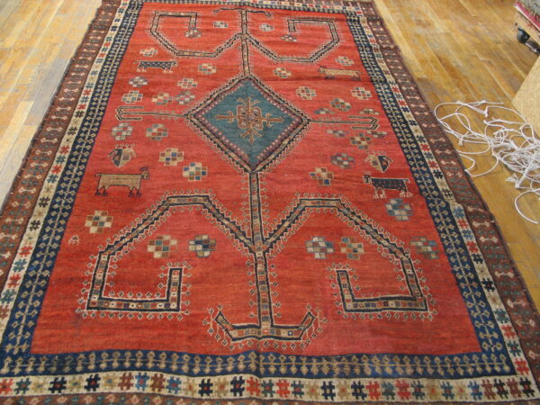 Rusty red tribal rug with a geometric pattern, central teal diamond medallion, and navy borders on wood floor.