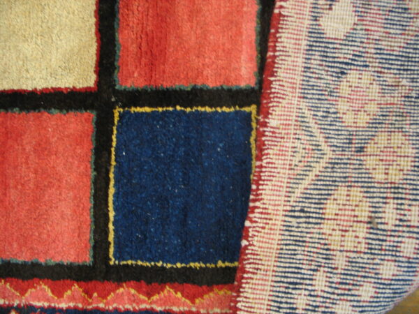 Geometric rug pattern of red, blue, and beige blocks with black borders, viewed beside the woven backing.