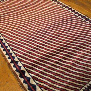 Red, cream, and dark blue striped rug with a zig-zag border rests on a wood floor.