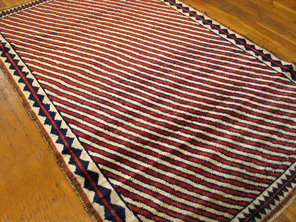 Red, cream, and dark blue striped rug with a zig-zag border rests on a wood floor.