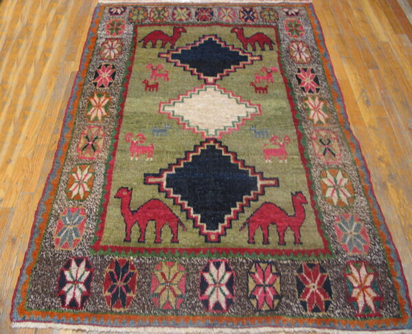 Traditional olive rug featuring stepped diamonds and red animals, framed by a colorful geometric border on wood.