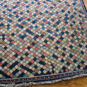 Medium pile rug with a variegated diamond pattern in blue, pink, cream, and yellow on a wood floor.